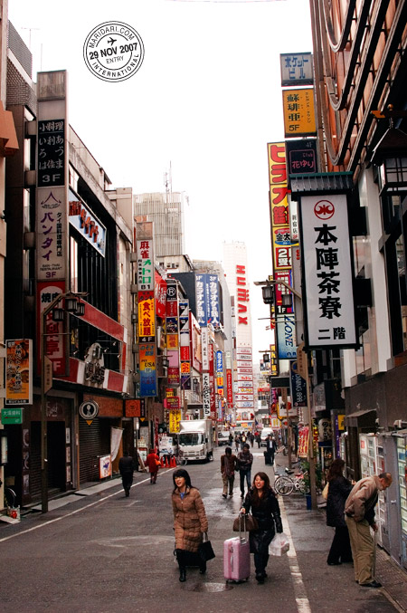 Tokyo street scene - Shinjuku neon signs in the day, two Japanese women with trolley bags